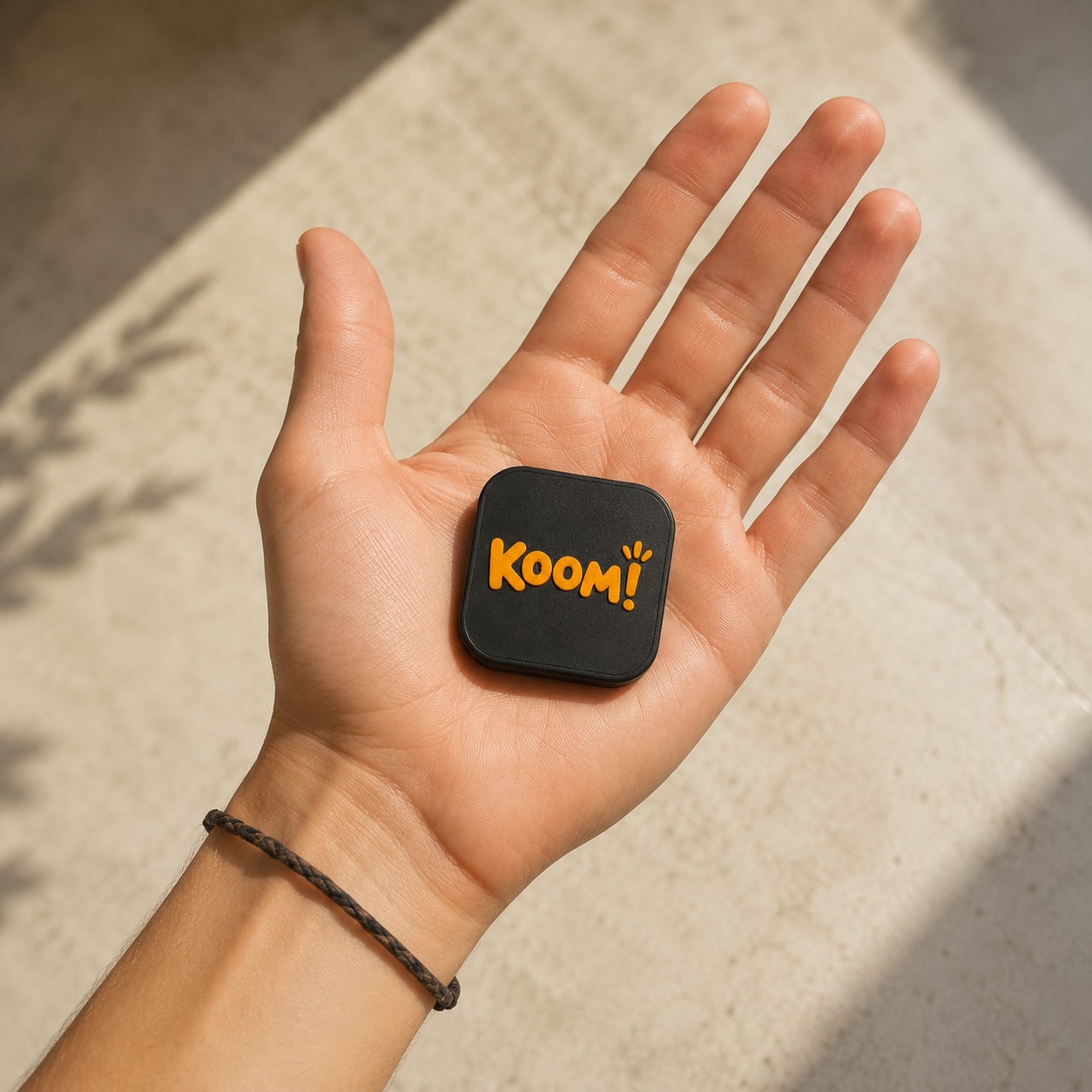 תג Koom!‎ מונח על כף יד - בערך בגודל של קופסת גפרורים